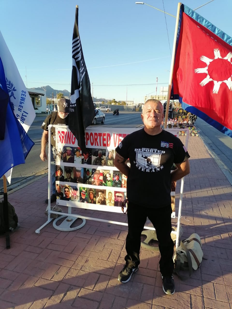 Marcelino Ramos, a 54 year old man, stands in front of a banner that has flags behind it and photos of deported veterans on the front. He is wearing a black tshirt and black pants.