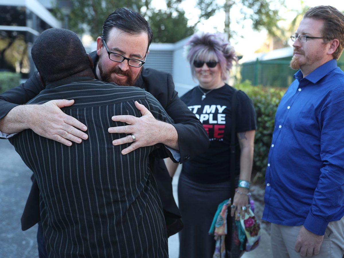 A white man with short brown hair, glasses and a beard and mustache hugs a Black man wearing a pinstripe suit whose back is to the camera, while a white woman with blonde hair and sunglasses and a white man in a blue, button-down shirt stand in the background wearing a partially obscured Black T-shirt that says, "Let My People Vote."
