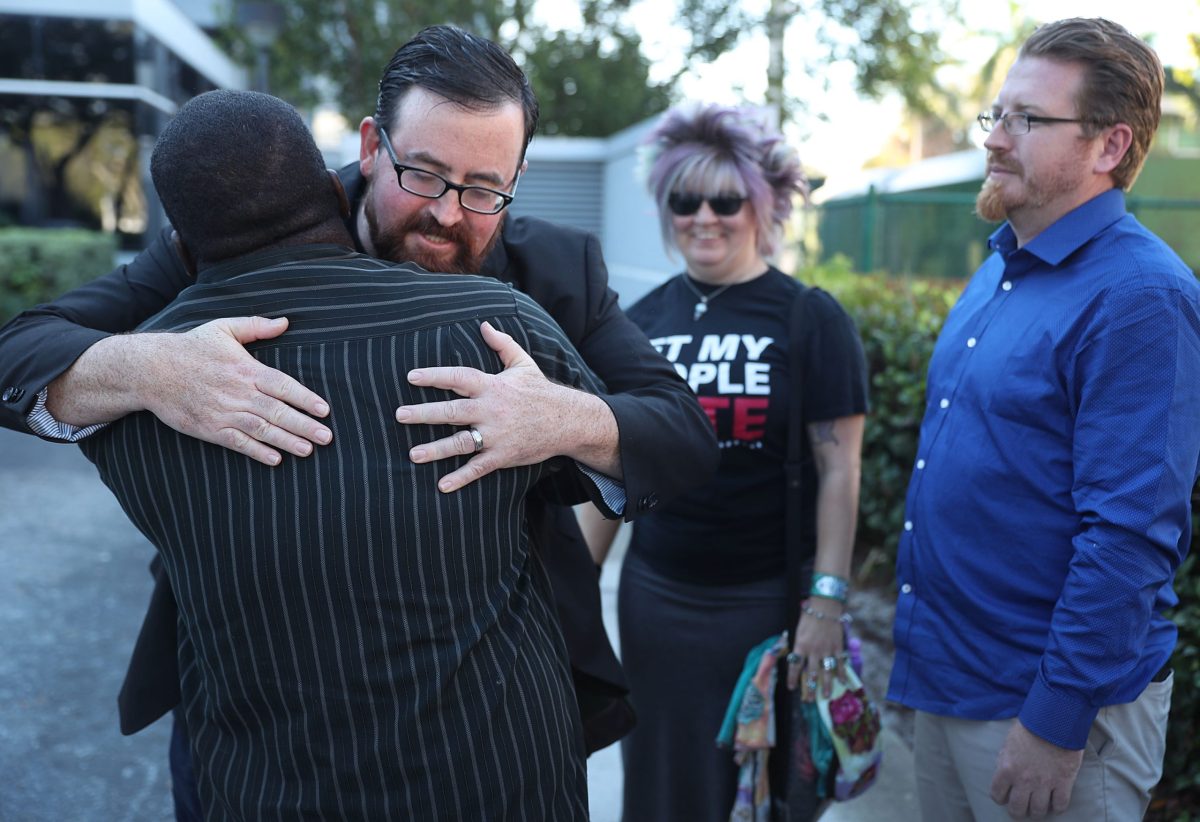 A white man with short brown hair, glasses and a beard and mustache hugs a Black man wearing a pinstripe suit whose back is to the camera, while a white woman with blonde hair and sunglasses and a white man in a blue, button-down shirt stand in the background wearing a partially obscured Black T-shirt that says, "Let My People Vote."