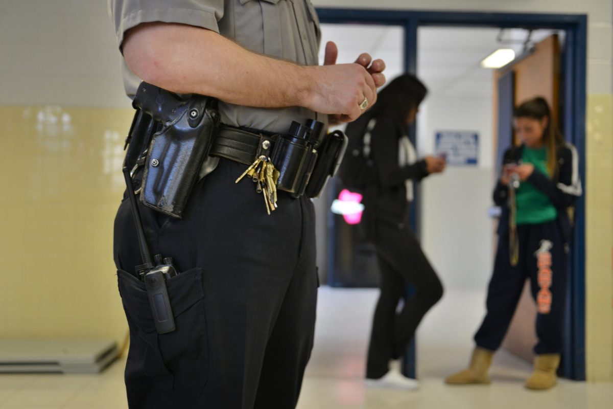 a police officer stands in a school hallway. Two students stand behind him out of focus. The viewer can see the items on his belt like keys, mace, and his holster.