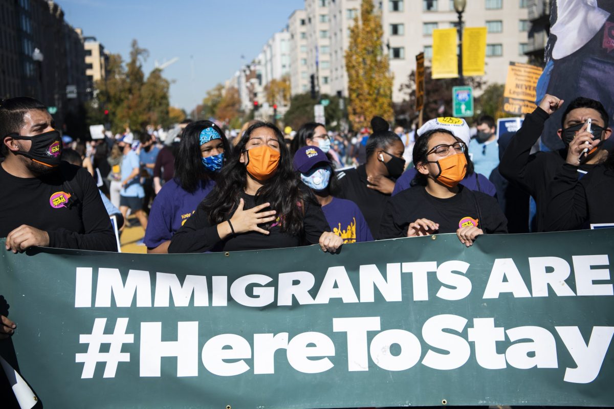A woman wearing an orange face masks holds her hand over her heart as she holds a green vinyl banner with other demonstrators that says Immigrants are #HereToStay. Other demonstrators are pictured behind them along with Washington, D.C., buildings.