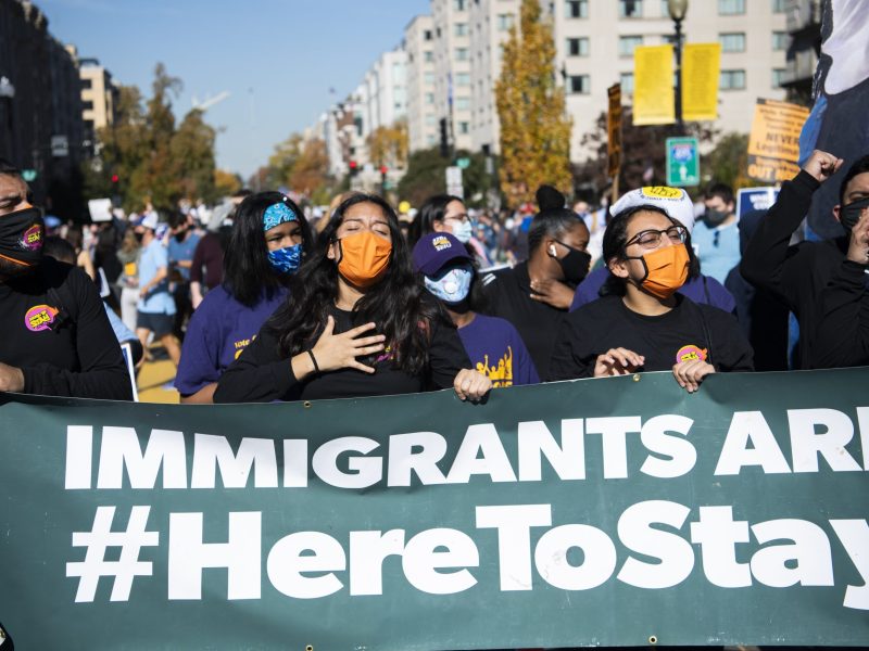 A woman wearing an orange face masks holds her hand over her heart as she holds a green vinyl banner with other demonstrators that says Immigrants are #HereToStay. Other demonstrators are pictured behind them along with Washington, D.C., buildings.
