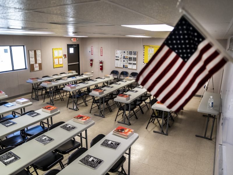 Text books and notebooks line tables inside a classroom while an American flag hangs in the front of the class.