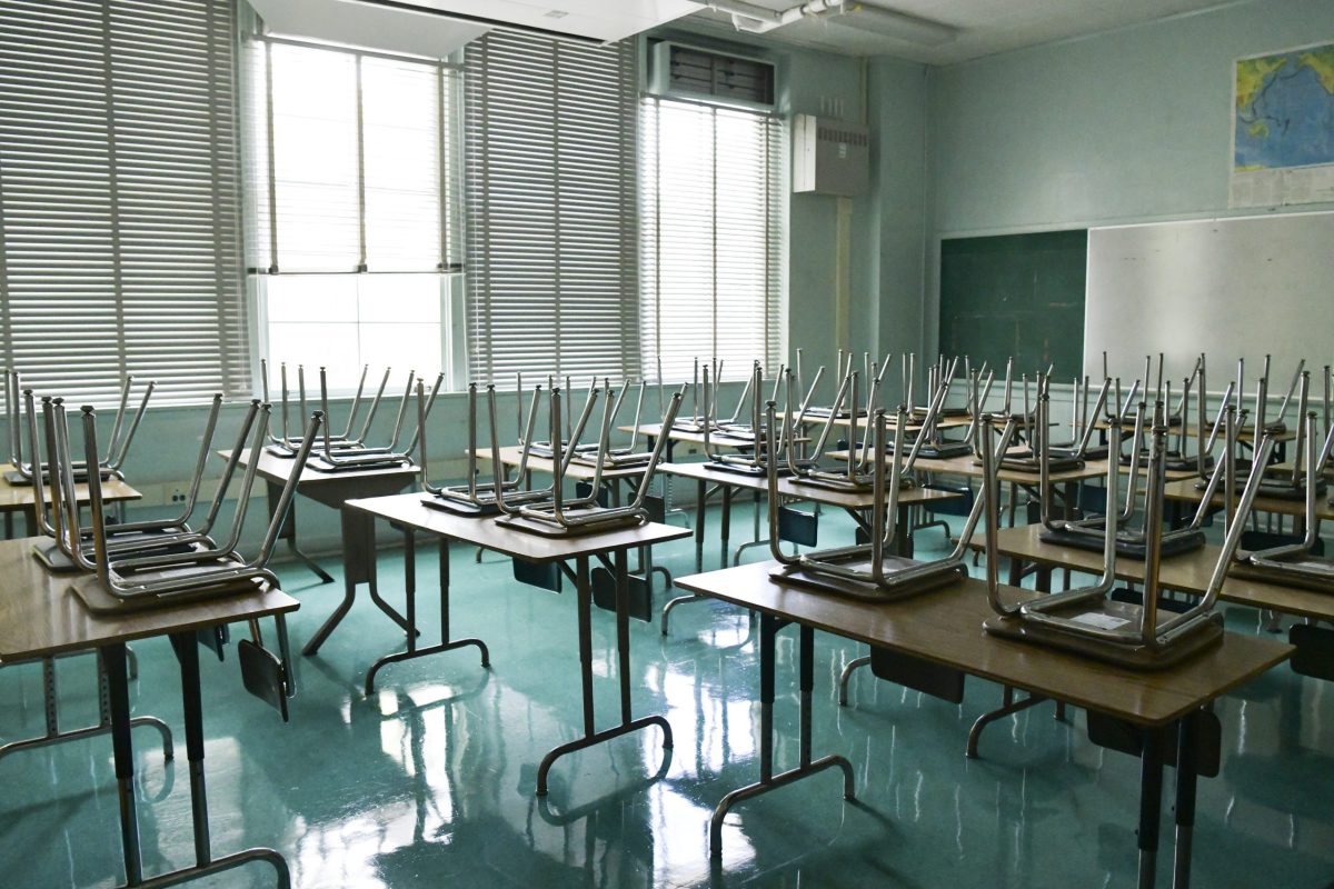 In an empty classroom, seats are upside down on desks as the sun shines in the windows.