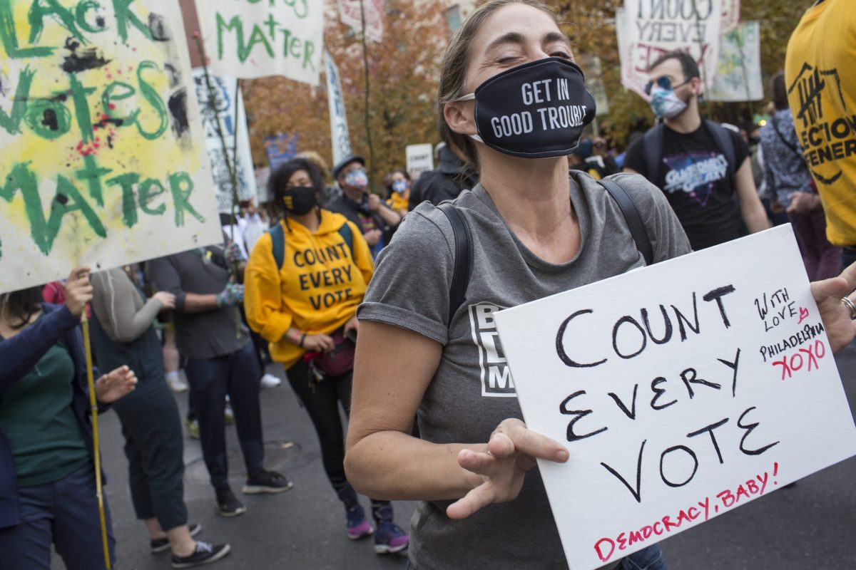 A woman standing in front of other protesters holding signs wears a gray shirt and a black face mask that says "Get in Good Trouble" on it and holds a white homemade cardboard sign that says "Count Every Vote" in black letters, with the words, "Democracy, Baby!" and "With love, Philadelphia XOXO" in smaller read letters.