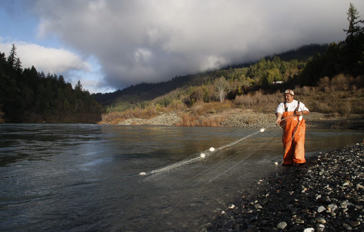 A Yurok tribe member wearing orange coveralls checks the net they have placed in a large body of water. there are small hills behind them and a cloudy sky.