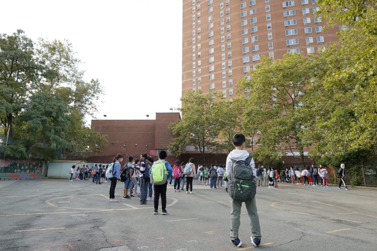 Students stand and walk along the blacktop toward red brick school buildings. There are green trees that line the way.