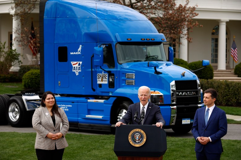 President Biden stands behind a podium as he speaks in front of the White House. Behind him is a tractor-trailer. 