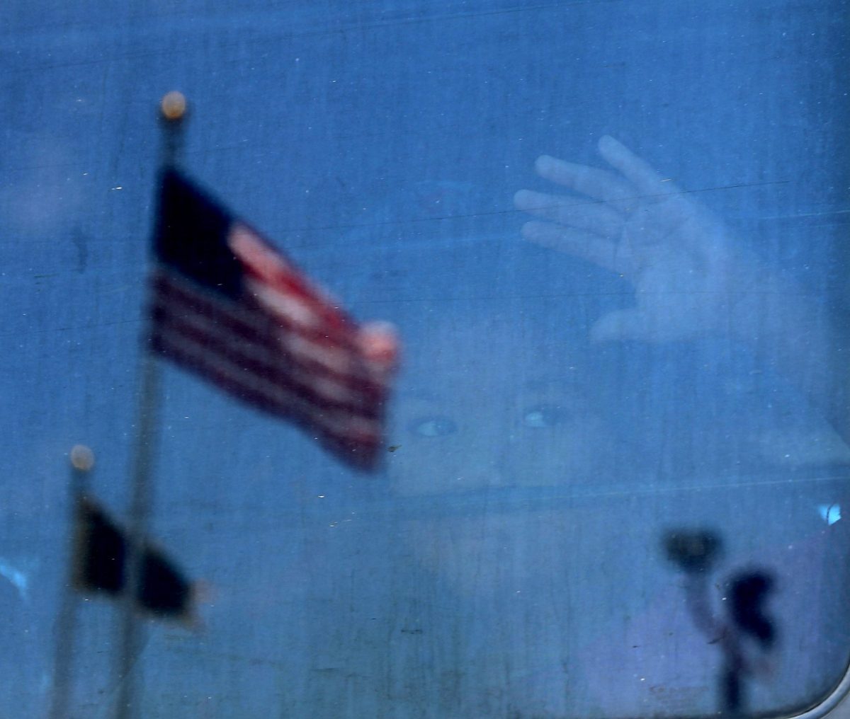 A child looks out of the window of a bus, in the reflection of the window you can see two American flags.