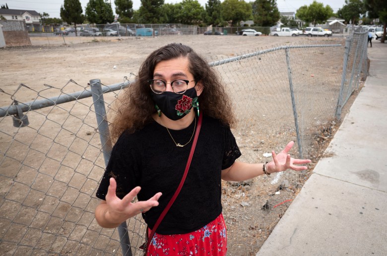 Patricia J. Flores Yrarrázaval gestures with her hands as she looks down the street with any empty lot behind her. She is wearing a Covid mask.