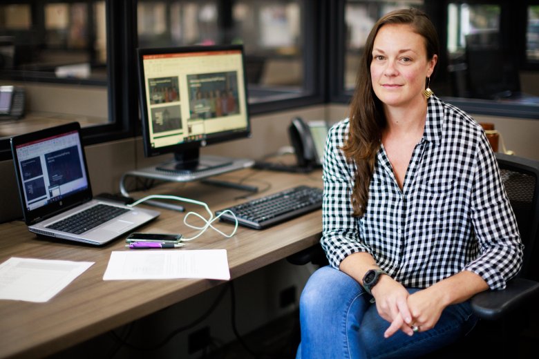Lorin Griffitts sits beside a computer. She's wearing a checkered shirt and jeans and has long brown hair.