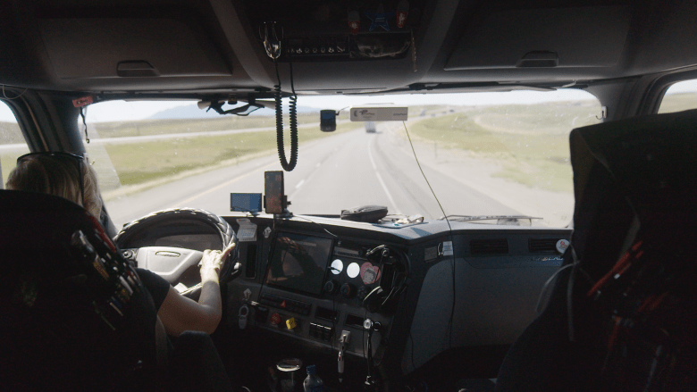 A view from the cabin of a tractor-railer driven by a female trucker. On the dashboard is a cell phone.